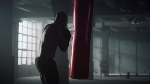 African american boxer hits punching bag during boxing training in dark gym
