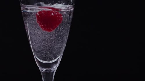 Cinematic highframerate cocktail shot. A raspberry falls inside glass of clear water and swirls up o
