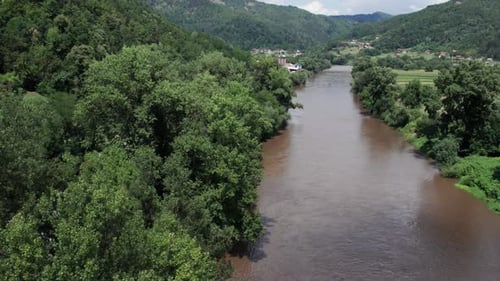 Drone view of muddy river surrounded by lush green forest and farmland