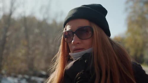 Une femme blanche en gros plan en écharpe et lunettes de soleil, rousse, exhale un souffle visible dans un parc froid subtil