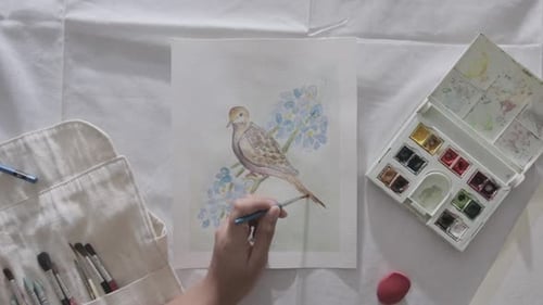 Caucasian left hand and fingers paints watercolor drawing of bird with brush on white paper, overhea