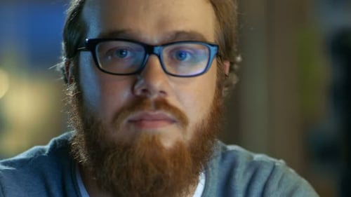 Young Bearded Creative Man With Glasses Thinks on a Problem While Working on His Personal Computer.