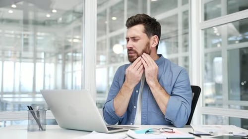 Upset businessman suffering from toothache working on laptop sitting at workplace at desk in busines