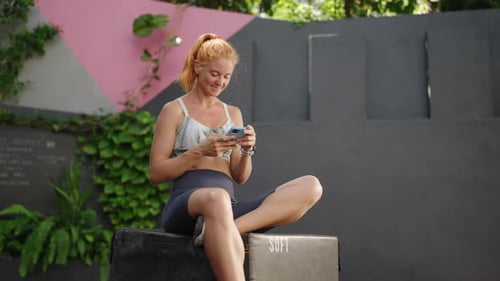 Redhead Female Athlete Checks Fitness App on Smartphone at Luxury Tropical Gym Sits on Bench