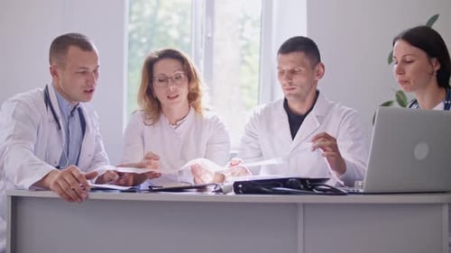 Team of Four Doctors in White Coats Sitting at Table in Hospital and Working With a Laptop