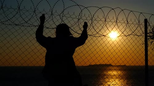 Refugee Silhouette Clinging to Barbed Wire Fence at Sunset