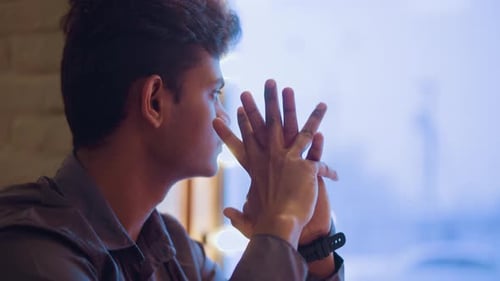 Young Man Looking Thoughtful Out Window, Hands Clasped