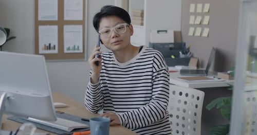 Confident Asian Businesswoman Speaking on Mobile Phone Discussing Work Sitting at Desk in Office