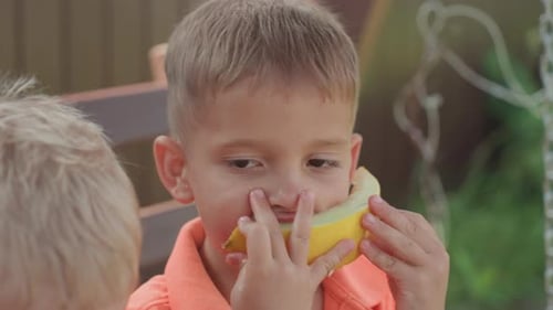 Enfant faisant un geste ludique avec une tranche de melon près du visage dans le jardin, garçon assis sur un banc utilisant du melon