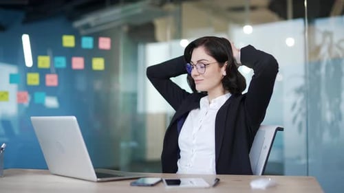 Businesswoman in office relaxing with hands behind head, sitting at desk with laptop.