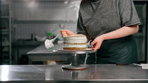 Close Up Female Baker in a Professional Close Up Kitchen Distributes Cream Onto Sponge Cake with a