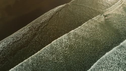 Aerial view of ocean waves rolling over dark sand beach