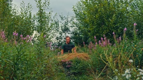 A Man is Pushing a Wheelbarrow Filled With Hay Through a Lush, Green Field Surrounded by Tall