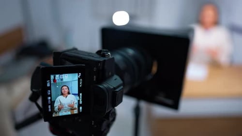 Camera recording a video with a long-haired Caucasian woman in white blouse sitting at table talking
