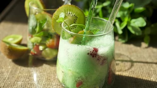 Pouring Foamy Soda Into a Glass with Pieces of Kiwi and Strawberries with a Leaf of Fresh Green Mint