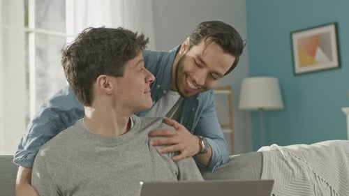 Sweet Male Gay Couple Spend Time at Home. Young Man Works on a Laptop, His Partner Comes From Behin