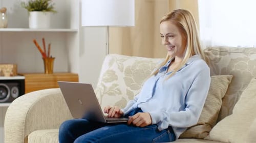 Long Shot of a Smiling Beautiful Woman Sitting on a Couch in Her Living Room. She Works on Her Lapt