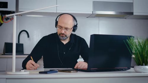 Professional Creative Man in Headset Sitting at Desk in Home Office Studio Working on Laptop Writes