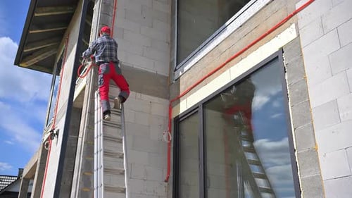 Worker Climbs Ladder to Paint Exterior of Building Under Clear Blue Sky