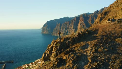 Man Backpacker Standing on Top Mountain Cliff with Rocky Beach and Ocean at Sunset
