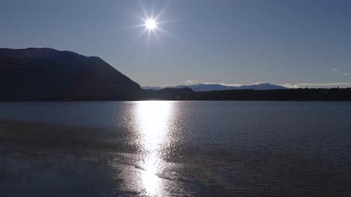 Sun reflecting on highland lake surface in aerial view at sunset with mountain silhouettes in