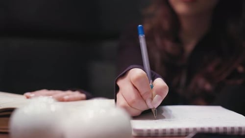 Close Up Female Hand Writing With Pen While Holding Open Book On Table