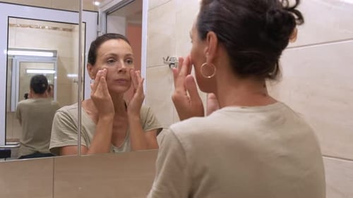Woman Applying Face Moisturizer in Bathroom Mirror