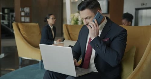 Ambitious Businessman Talking on Mobile Phone and Typing with Laptop in Hotel Lobby