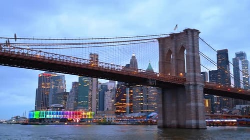 Approaching the Brooklyn Bridge by the river. Luminous New York skyline at backdrop.