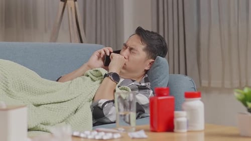 Sick Asian Man With Blanket Suffering From Cough While Talking On Phone With Doctor Lying On Sofa