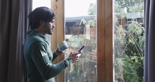 Caucasian man holding cup of coffee using smartphone by window at home, copy space, slow motion