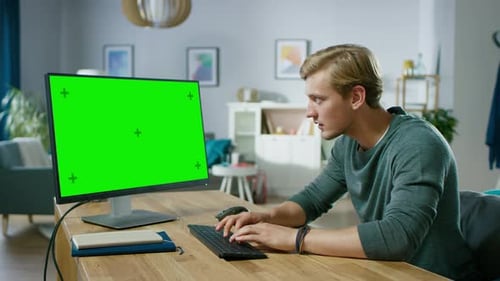Handsome Young Man Works on a Green Mock-up Screen Personal Computer while Sitting at His Desk in t