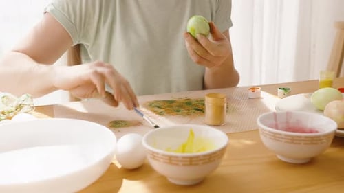 Woman working on a green Easter egg in a bright sunny room, with paint bowls and brushes scattered o