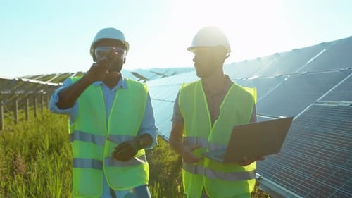 Young Two Men Use Laptop on the Solar Farm in Special Uniform Walking Discuss the Installation of