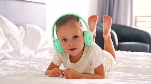 Little Girl Laying in Bed Singing and Listening Music with Green Kids Headphones in Home Bedroom