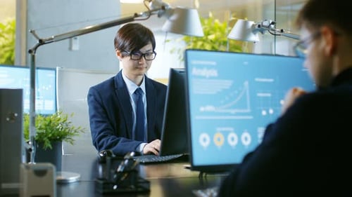 In the Office East Asian Businessman works on a Desktop Personal Computer. His Colleague's Computer
