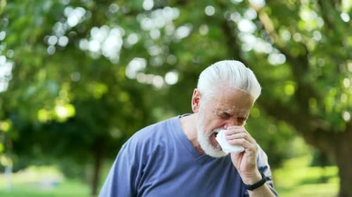 Senior active gray haired bearded man suffering from seasonal allergies standing in urban city park.
