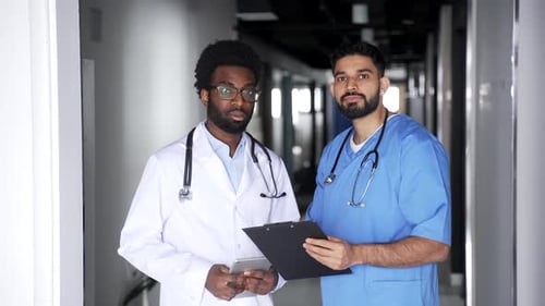 Two confident serious doctors in coats standing in hospital corridor holding clipboard and tablet.