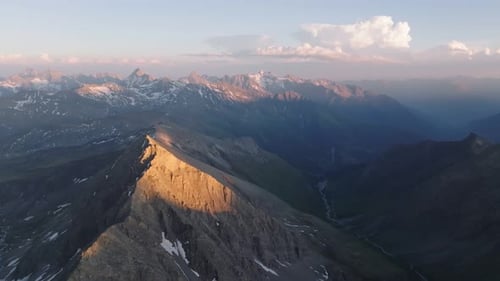 Dramatic drone aerial of snow-flecked alpine ridge at sunset, descending into a deep mountain valley