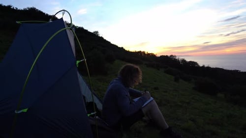 Journaling man writing notebook in nature ocean campsite at sunset