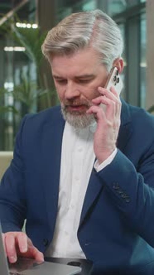 Mature Businessman in Suit Answering to Client on Mobile Phone Call Using Laptop in Office Lobby