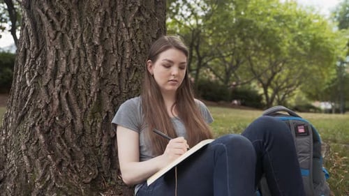 Caucasian College Girl Writing In Journal Sitting Under A Tree At Campus Park On Beautiful Day