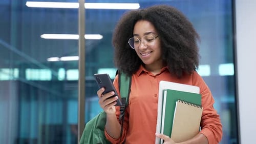 Smiling african american female student is using mobile phone standing at university indoors. Happy
