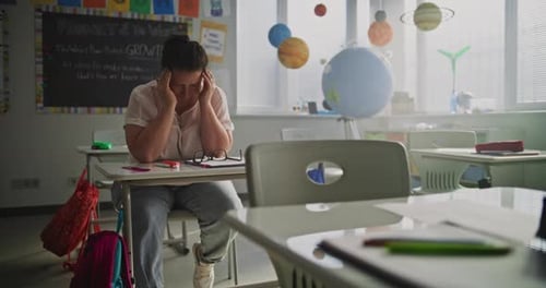 Exhausted Female Teacher Rubbing Her Temples Sitting Alone at Desk in Empty Classroom