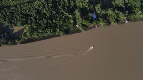 Aerial top down shot of boat cruising on brown colored amazon river and arriving dock in forest. Hig