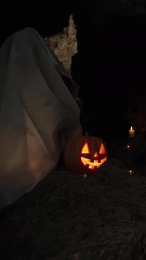 Vertical video. A Ghost in a White Sheet Sits in a Dark Cave on Halloween Night.