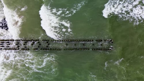 Aerial view of ocean waves crashing against rows of stone breakwaters. The geometric pattern