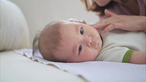 A tiny child smiles happily on the bed and mother is using a cotton bud to clean child ears