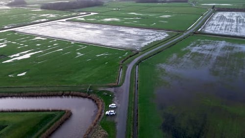 Luftaufnahmen von überfluteten grünen Feldern, die von engen Straßen und Kanälen durchschnitten werden. Zwei weiße Autos