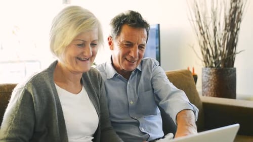 Senior Couple Smiles While Using Laptop At Home